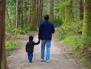 Back side of father and son holding hands while walking down a wooded path