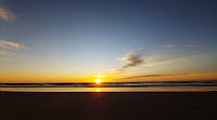 Bright sun rising on the horizon of a heavily shaded beach and ocean in the lower third of the image and a deep blue sky partically covered with orangeness from the sun and a few streaks of cloudswith orange coloring from the sun and a few clouds to the sidewith sun rising at the horizon