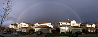 Full rainbow above neighborhood houses against an overcast sky. Garbage bins are in front of the houses