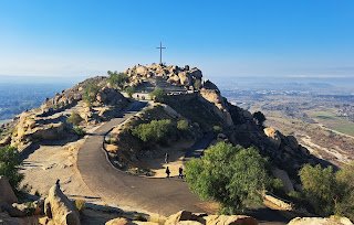 Atop a mountain a curved concrete path circles it's way toward a cross that stands high above everything else against a clear blue sky