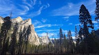 Rich blue sky with silky swirls of clouds set behind a massive, granite mountainscape with a hint of a meadow in the foreground surrounded by thin, tall trees reaching for the sky